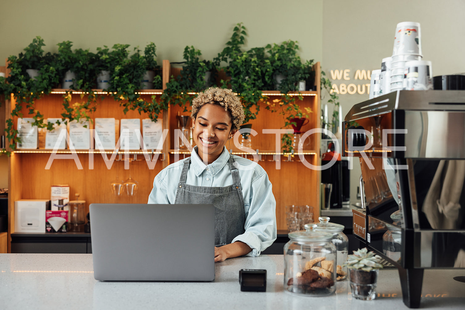 Young smiling woman in an apron typing on a laptop. Female working as a barista standing at the counter with a laptop.