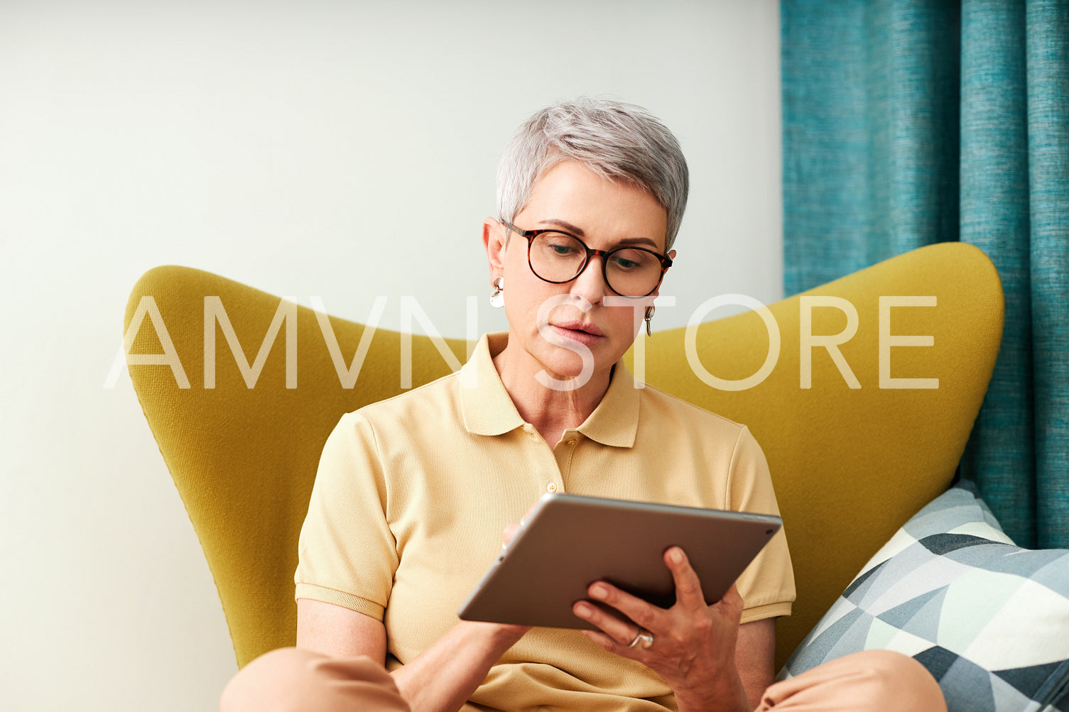 Mature woman sitting at home on armchair using digital tablet. Female in casuals watching content on a tablet.	