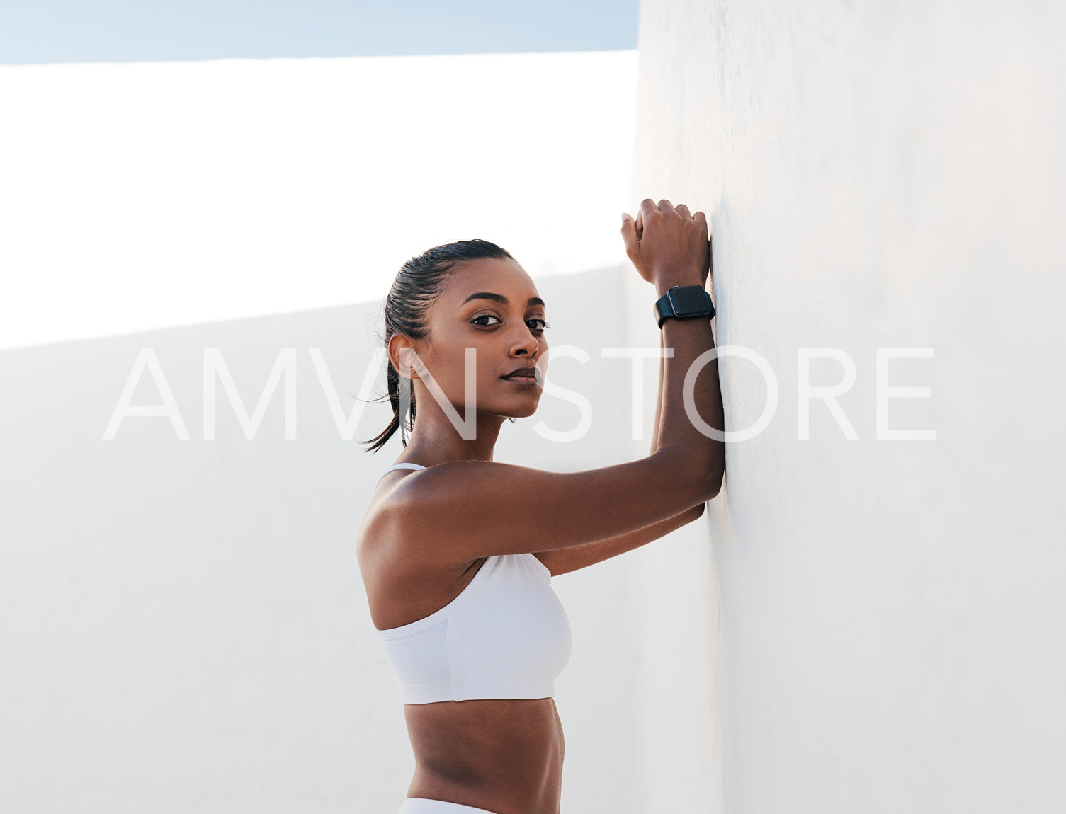 Confident slim female relaxing at the wall. Young fitness influencer in sports bra looking at camera.