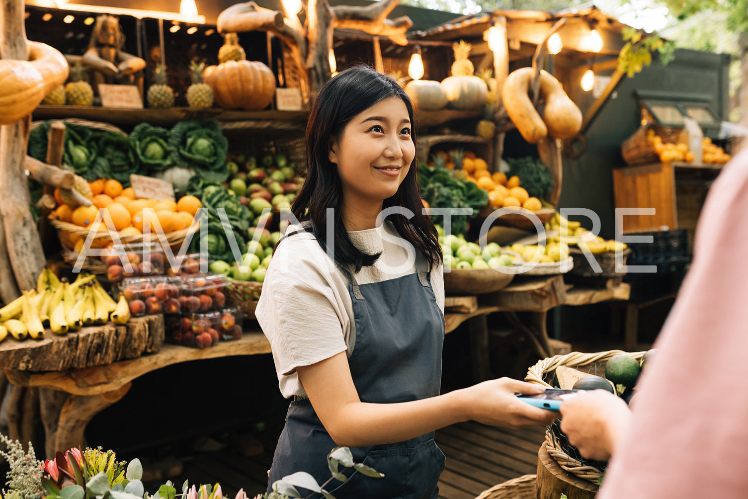 Outdoor market owner receiving payment from the buyer. Asian woman in an apron holding a pos terminal looking at a customer at a local farmer market.