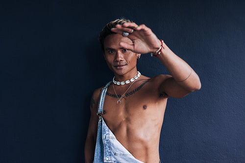 Guy with a bare chest hiding his face with one hand while standing at a blue wall outdoors