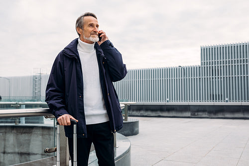 Mature male tourist standing outdoors with luggage and talking on cell phone