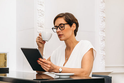 Mid adult woman holding a cup and looking on digital tablet. Businesswoman reading while having breakfast.