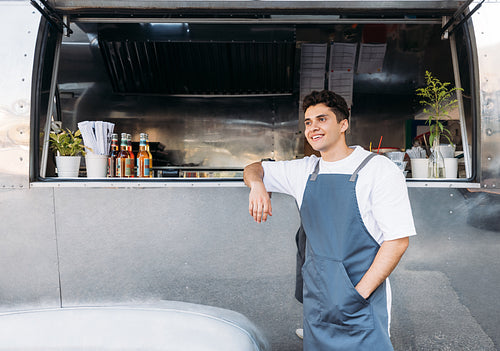 Young business owner in an apron. Salesman leaning on a food truck.