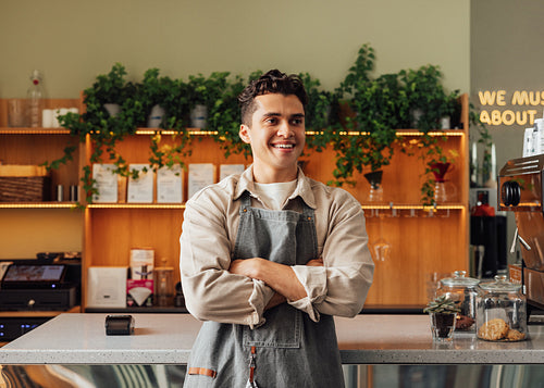 Confident barista in an apron. Young guy working as a barista in a coffee shop standing with crossed hands and looking away.