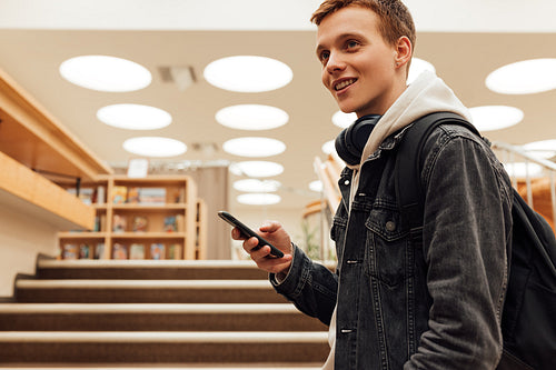 Smiling guy with smartphone standing in college library and looking away