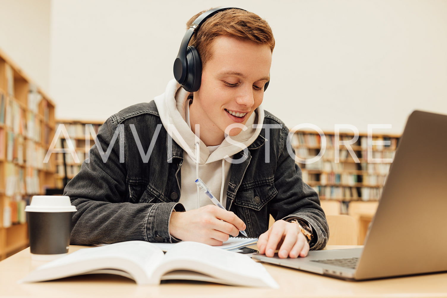 Smiling college student wearing wireless headphones while writing on exercise book in library. Guy with ginger hair doing homework.