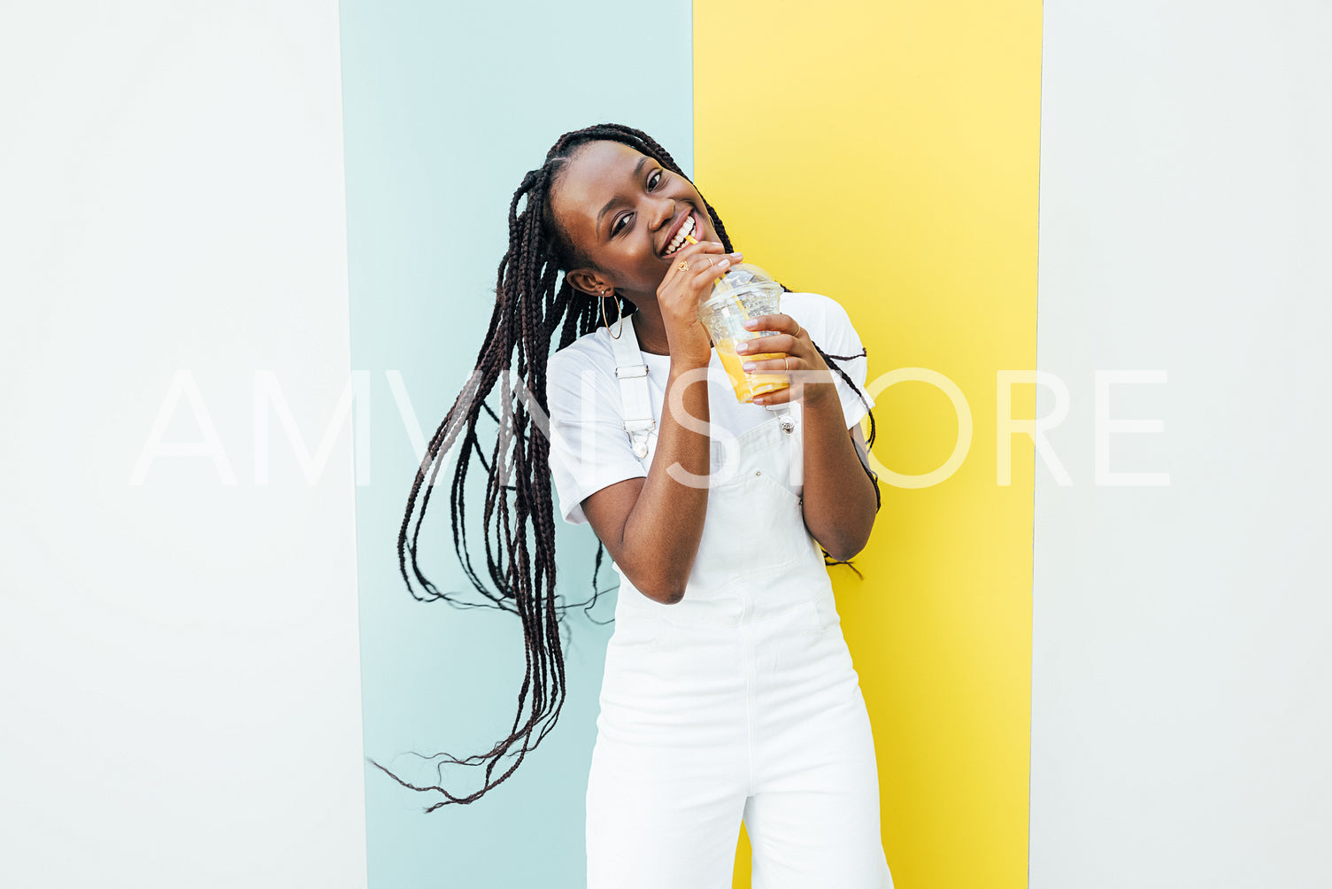 Playful woman in white overall drinking juice and looking at camera while standing at wall outdoors