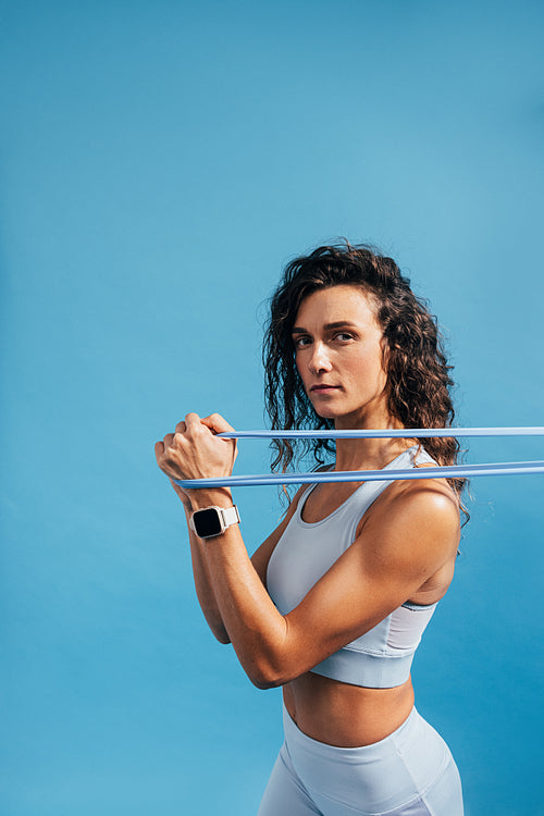 Sportswoman working out with elastic band on blue background background and looking at camera