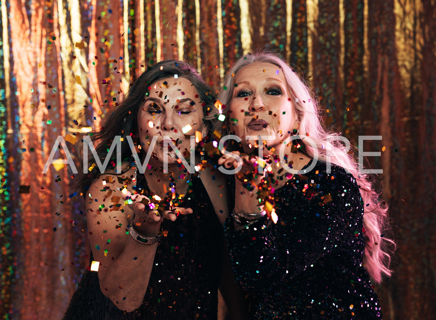 Two fashionable senior women in black dresses blowing confetti off their hands in studio