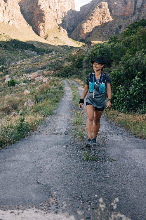 Smiling woman on hike. Female in sportswear walking on a road in valley and looking away.