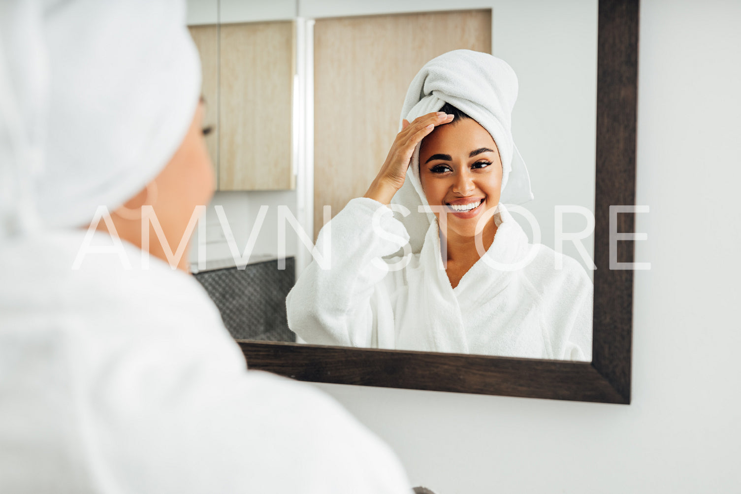 Beautiful smiling woman in bathrobe enjoying a spa day at home. Young female with a white towel on her head admiring her face in the mirror.