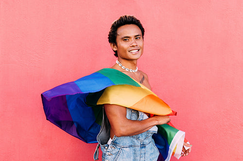 Portrait of a young smiling guy posing with LGBT flag against pink wall outdoors