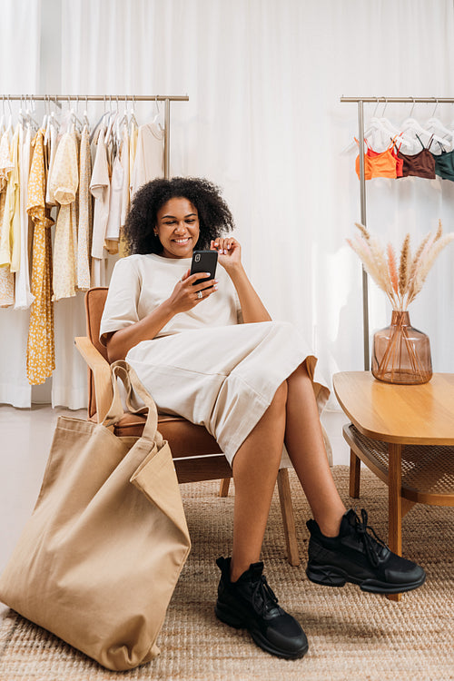 Smiling woman with mobile phone sitting on armchair in a clothing store. Customer relaxing in a boutique.