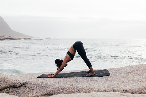 Side view of slim woman practicing Downward Facing Dog pose by ocean at sunset