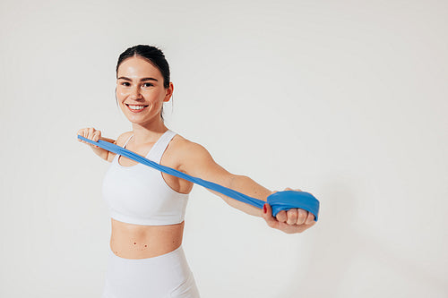 Cheerful woman in white fitness wear is warming up her arms with a resistance band in the studio