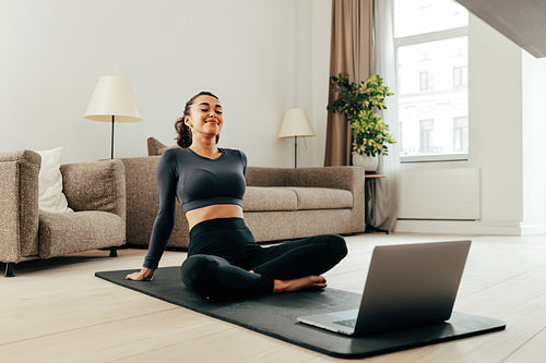 Smiling woman in sport clothes sitting on a mat in living room in front of a laptop. Young female relaxing during training at home.