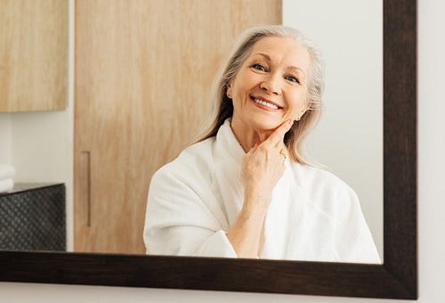 Portrait of an aged woman in a bathroom. Smiling woman with grey hair looking at her reflection in a mirror.