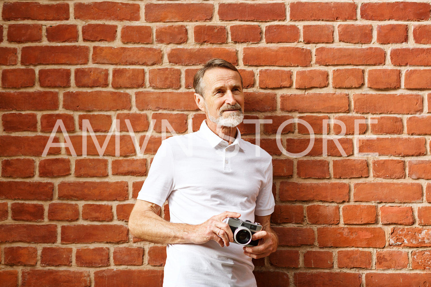 Bearded senior man standing with a film camera at a brick wall	