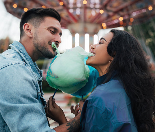 Young happy couple biting cotton candy. Man and his girlfriend bite a blue cotton candy against the carousel.