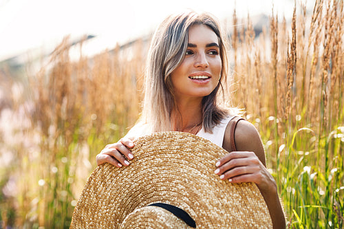 Young beautiful woman holding a big straw hat on a field surrounded by tall grass