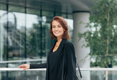 Portrait of a cheerful middle-aged female in black formal clothes. Businesswoman with ginger hair at railing outdoors.