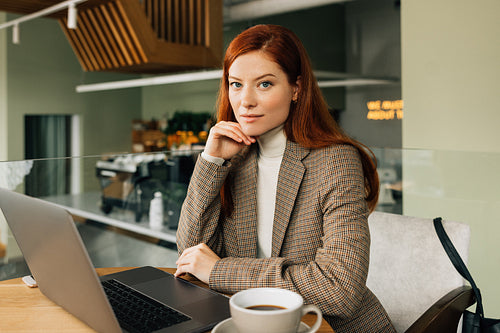 Portrait of a young businesswoman with ginger hair. Stylish female sitting at a table in a cafe with a laptop and coffee.