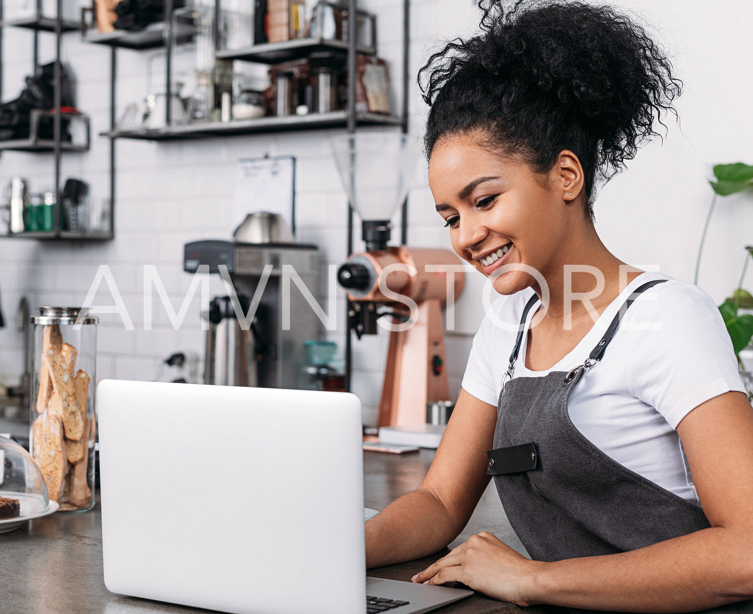 Smiling woman with curly hair wearing an apron typing on a laptop in a coffee shop