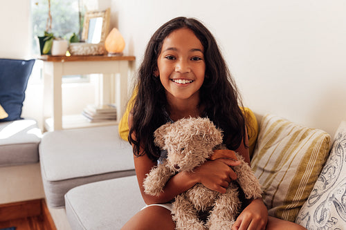 Portrait of a girl with a teddy bear looking at camera while sitting at home. Smiling kid with a toy.