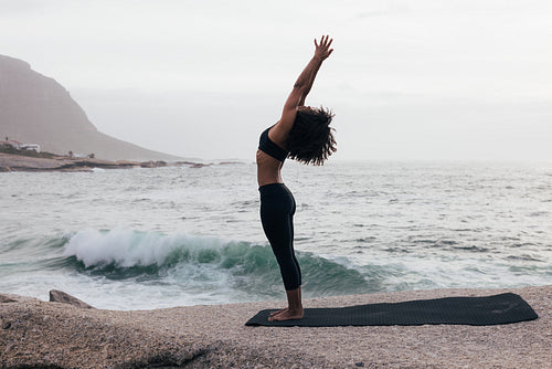 Side view of a slim female raising hands in yoga pose standing against an ocean at evening