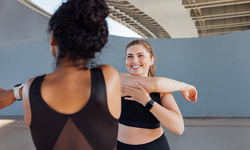 Plus size female smiling and looking at her fitness buddy while stretching her arm. Two women in black fitness attire warming up hands before a workout.