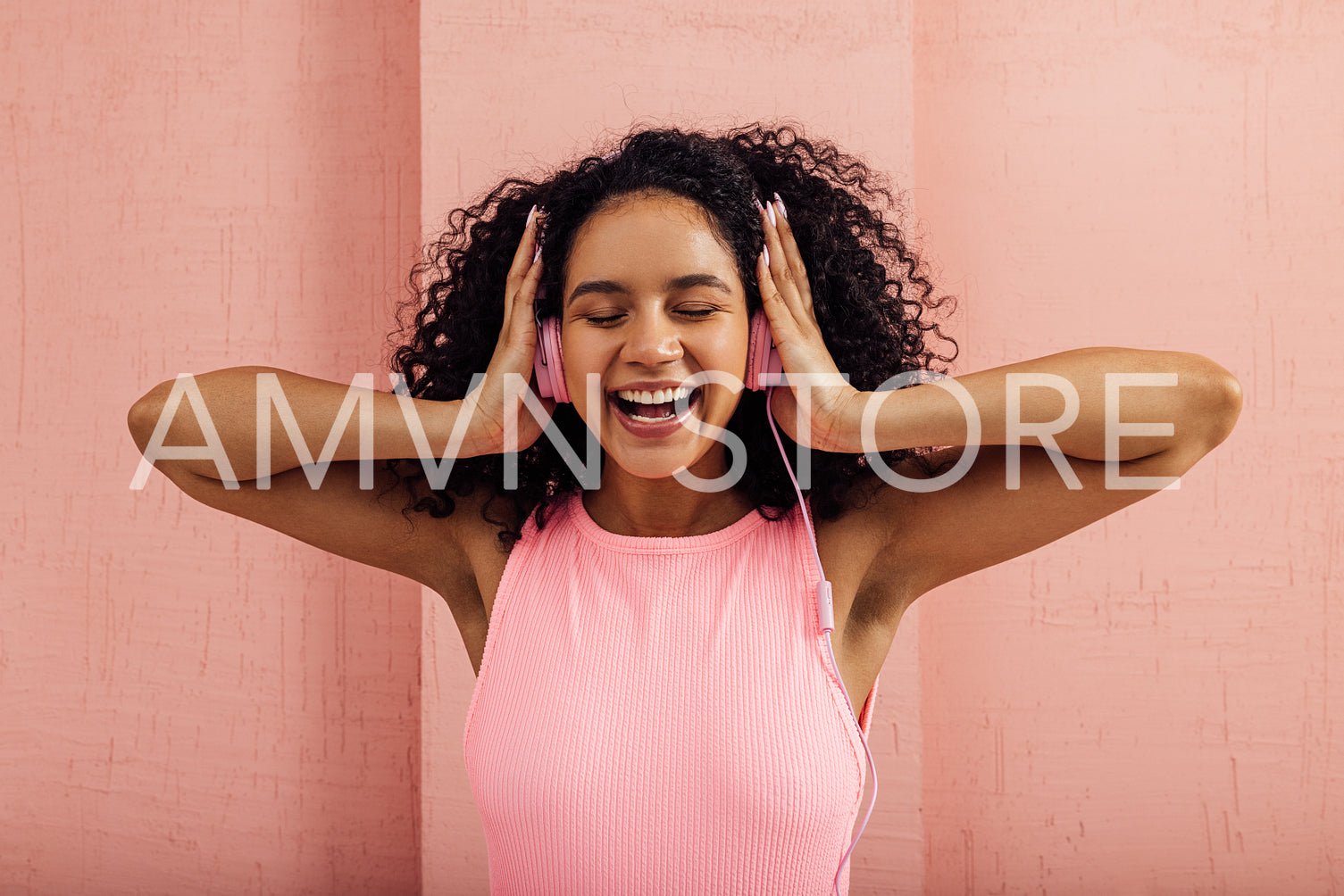 Laughing woman wearing pink headphones enjoying music with closed eyes. Smiling female with curly hair against a pink wall listening to music.