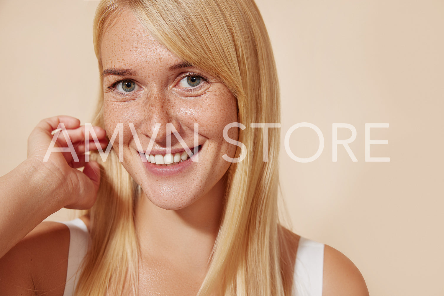 Close-up of a young freckled female looking straight at a camera in the studio. Female with perfect skin adjusting her long blond hair.