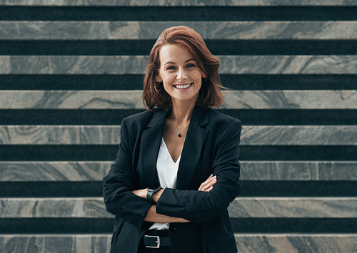 Happy female with ginger hair in formal wear looking at camera. Young confident middle-aged woman with crossed arms standing outdoors and smiling.