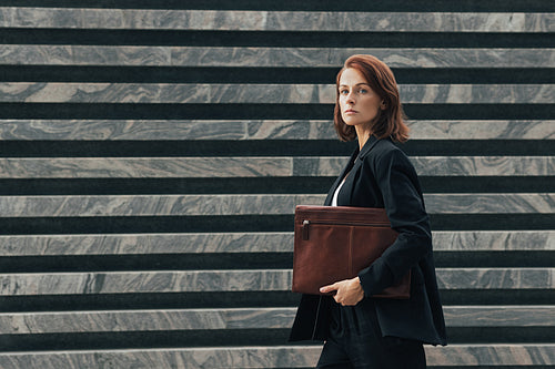 Side view of a confident businesswoman with a leather folder walking outdoors. Middle-aged businesswoman in black formal clothes looking away while walking.