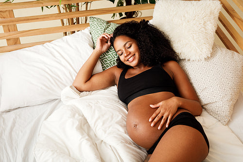 Smiling pregnant woman lying on bed at home, touching her belly