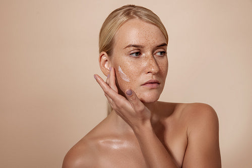 Confident woman with freckles applying anti aging cream on a cheek in studio while looking away