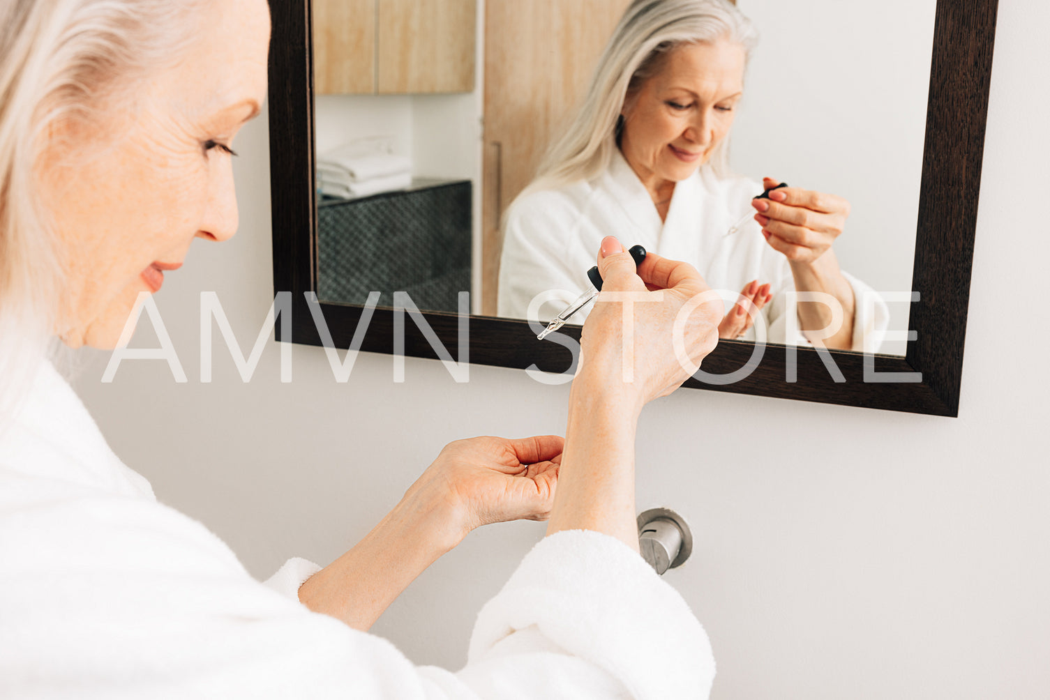 Senior female in a bathrobe drips liquid facial treatment on a palm in front of a mirror. Hands of an aged woman with a hyaluronic dropper.