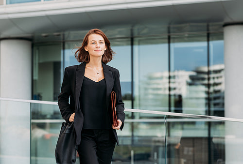 Confident female with a bag and leather clothes walking outdoors against a glass building. Cheerful corporate person walking outdoors.