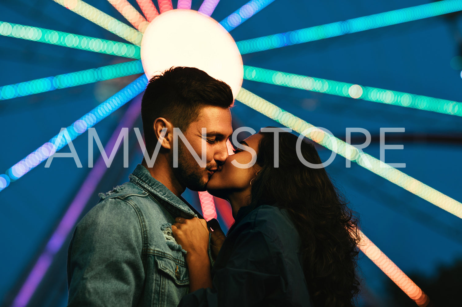 Young couple kissing in front of a Ferris wheel. Girl kissing her boyfriend in an amusement park at night.