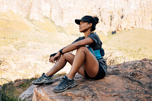 Side view of sportswoman sitting on a rock and relaxing. Young female hiker taking a break during exercises enjoying the view.