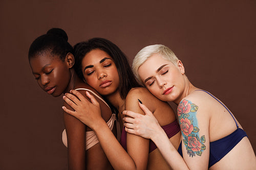 Three women in lingerie with closed eyes against a brown backdrop. Diverse females are embracing together.