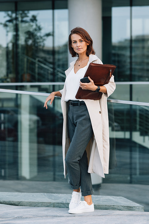 Confident middle-aged female holding a leather folder looking at camera standing at an office building