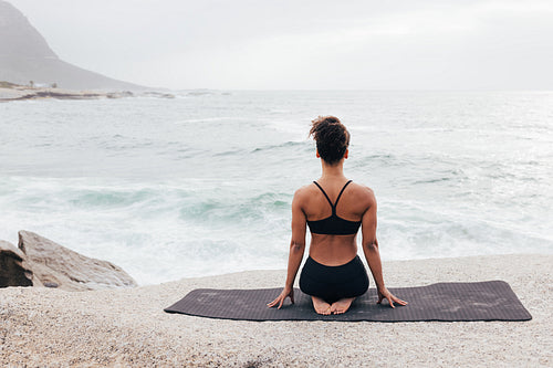 Woman sitting on a yoga mat and looking at ocean. Rear view of fit female sitting in Vajrasana pose outdoors.