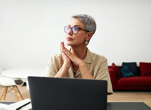 Stylish mature woman in glasses sitting at desk looking away