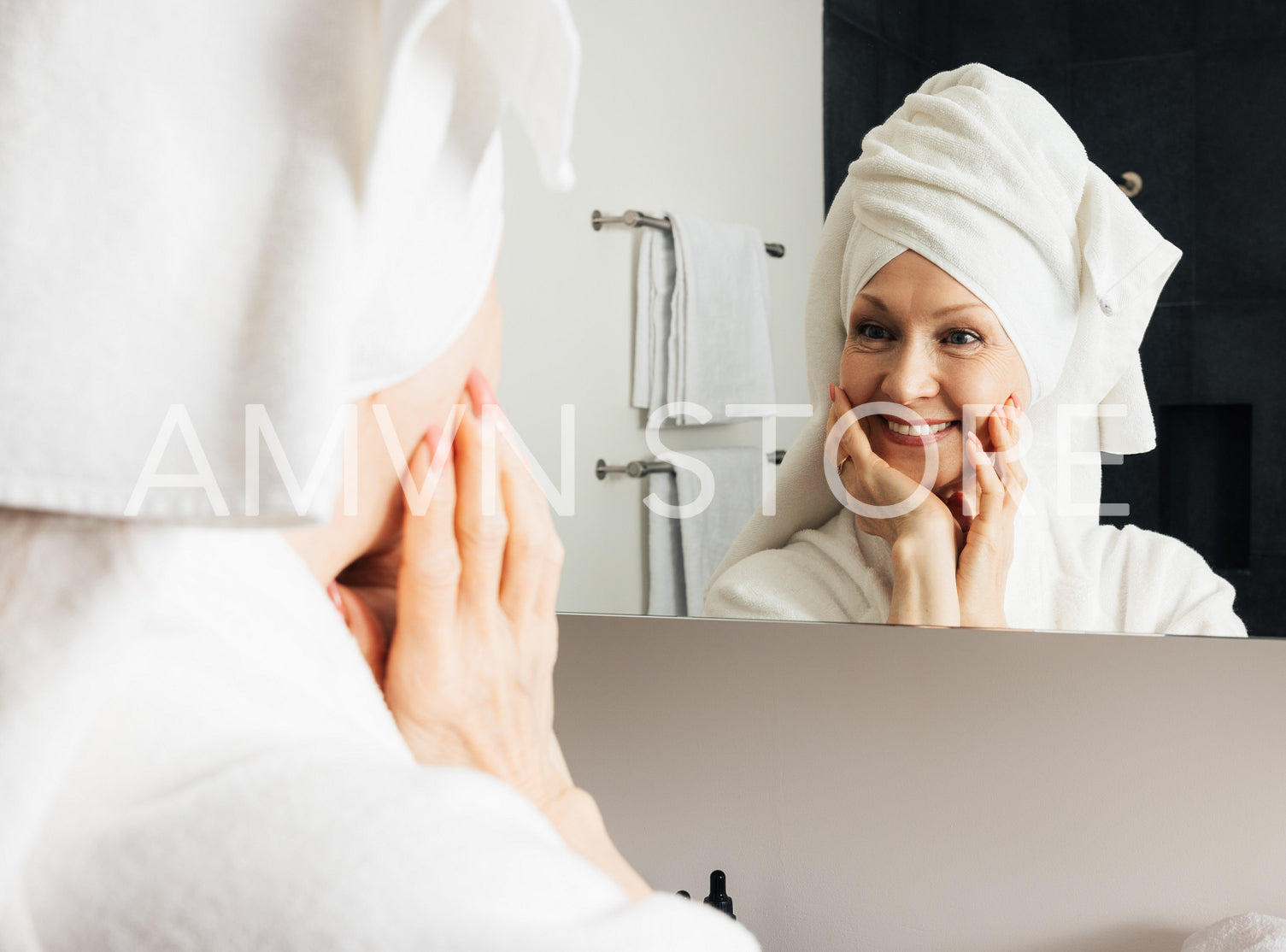 Cheerful woman admires her reflection in the bathroom mirror. Mature female touching her face with hands after the home spa.