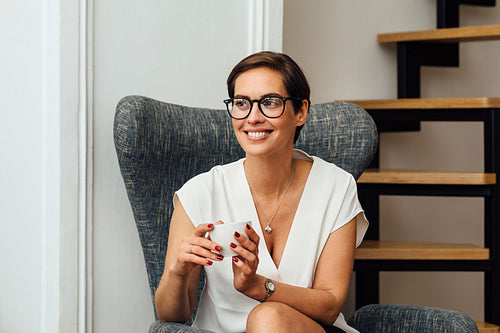 Smiling woman in eyeglasses holding a cup and looking away while sitting in apartment on an armchair