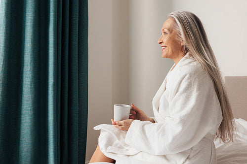 Senior woman sitting on bed in bedroom and looking away. Side view of pensive mature female.