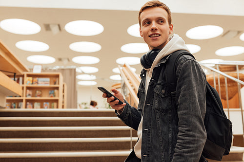 University student with bag standing in library. Teenage student holding smartphone and looking away.