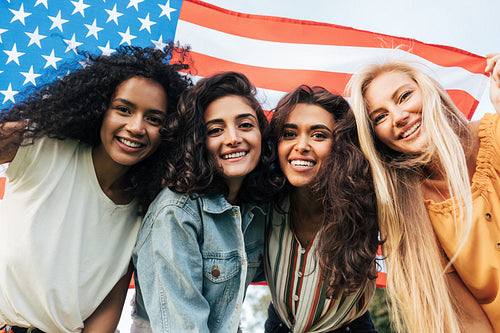 Four diverse women friends looking at the camera under the American flag. Cheerful females celebrating the 4th of July outdoors.